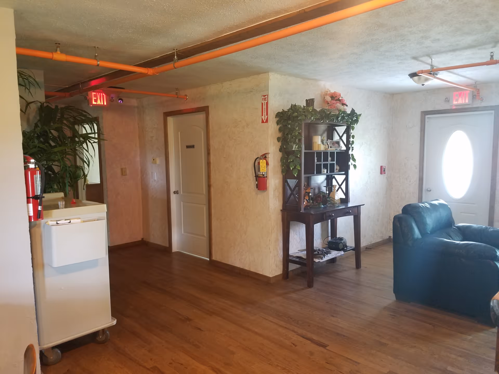 Interior view of a hallway in an assisted living facility with wooden flooring, beige walls, and orange exposed ceiling pipes. There is a white door, a fire extinguisher mounted on the wall, a wooden shelf with plants and decorative items, a green leather armchair, and two illuminated exit signs above doors.