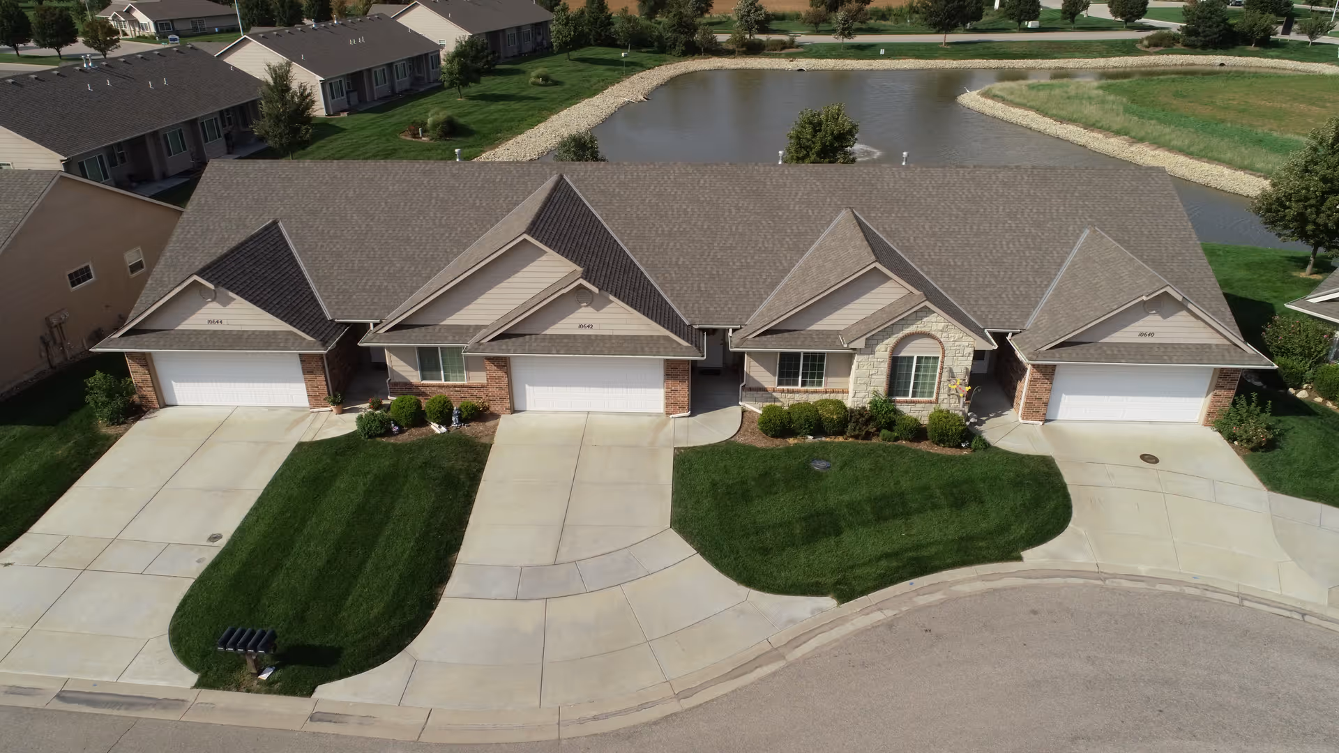 Aerial view of a row of single-story residential units with attached garages, well-maintained green lawns, and a small pond behind the buildings. The houses have gray shingled roofs and brick and stone facades.