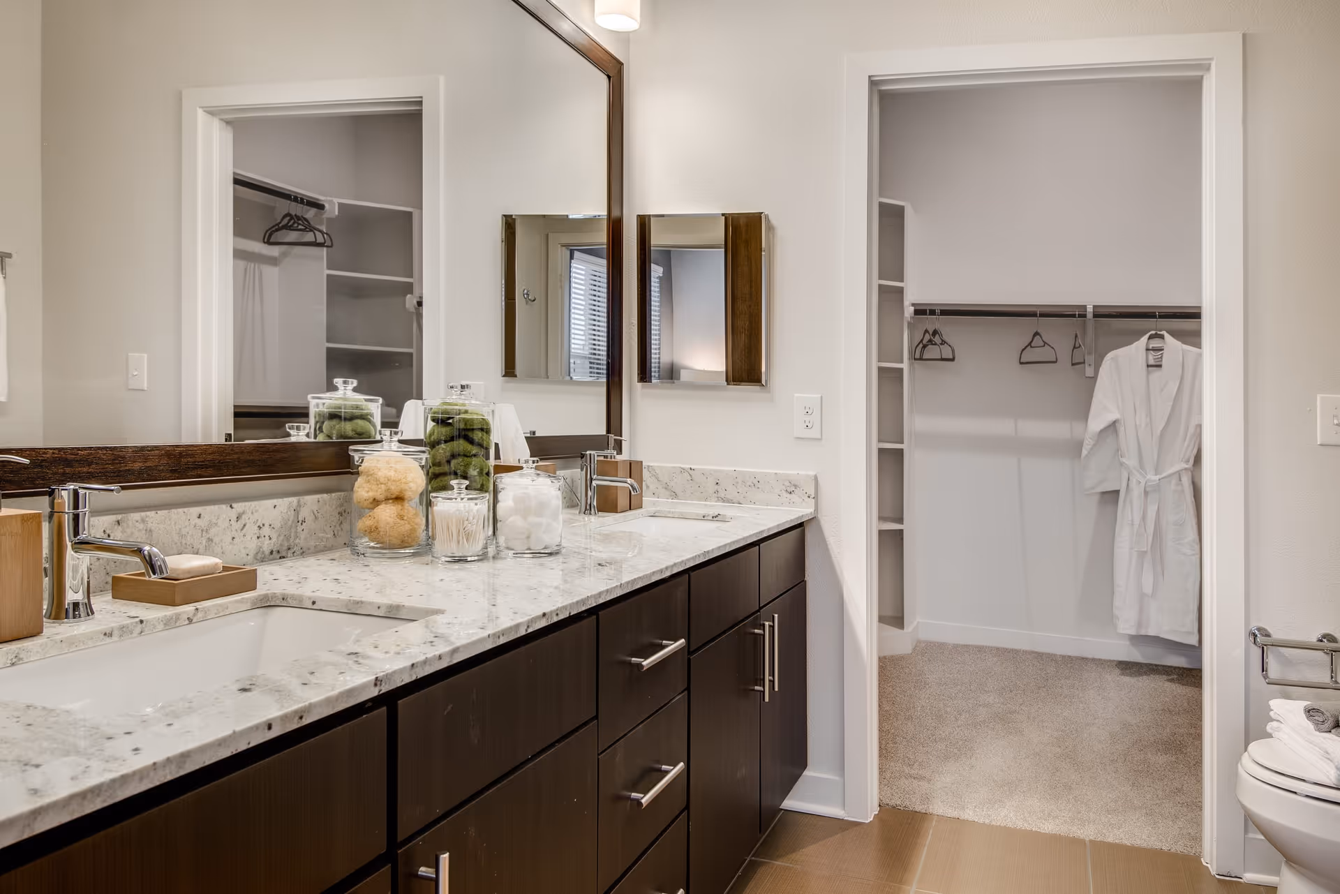 Modern bathroom with a granite countertop featuring two sinks, a large mirror, and jars containing bath items. A doorway leads to a walk-in closet with shelves, hangers, and a white bathrobe hanging inside.