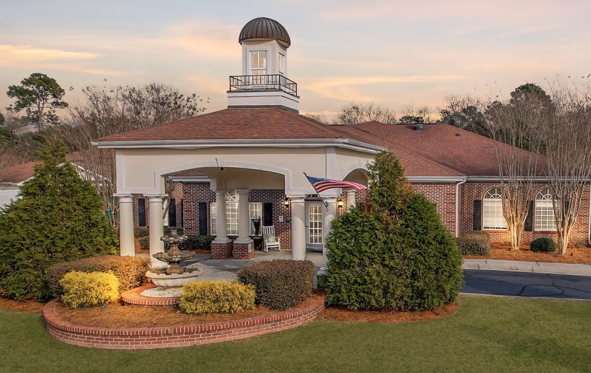 Exterior view of a senior living facility building with a covered entrance supported by white columns, a small fountain surrounded by bushes and greenery, an American flag, and a cupola on the roof. The building has red brick walls and multiple windows with black shutters.