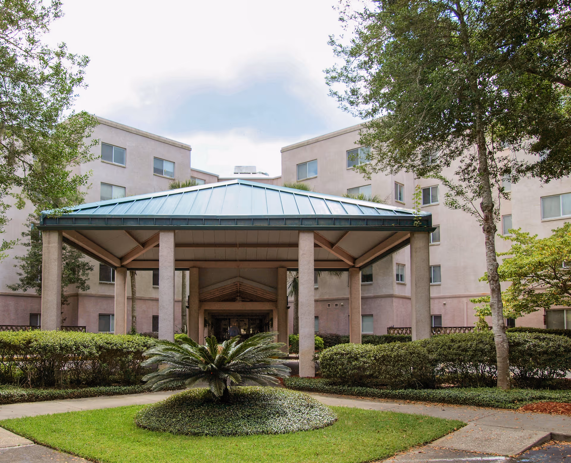 Covered entrance canopy with columns leading to a multi-story building surrounded by shrubs and trees.