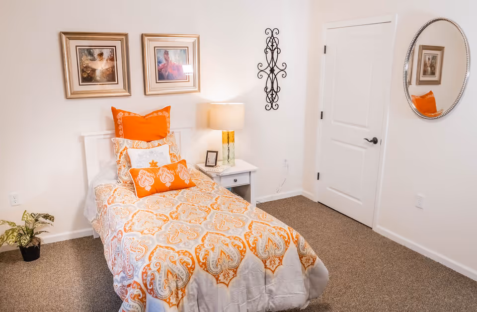 A neatly made single bed with orange and white patterned bedding and multiple orange and white pillows in a bright bedroom. There is a white nightstand with a lamp and a small framed picture next to the bed. Two framed pictures hang on the wall above the bed, and a decorative black metal wall piece is mounted on the adjacent wall. A round mirror is hung on the wall near a closed white door, and a small potted plant is on the floor near the bed.