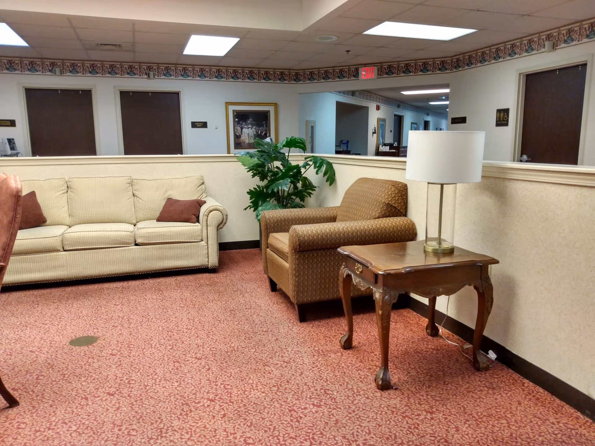 A seating area in an assisted living facility with a beige couch, a brown armchair, a wooden side table with a lamp, a potted plant, and a hallway with several closed doors in the background.