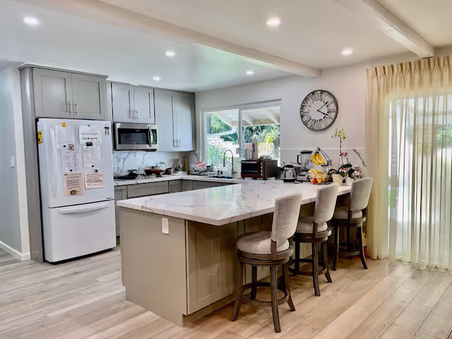 Modern open kitchen with a large marble-topped island, four upholstered bar stools, stainless appliances, and a sliding glass door letting in light.