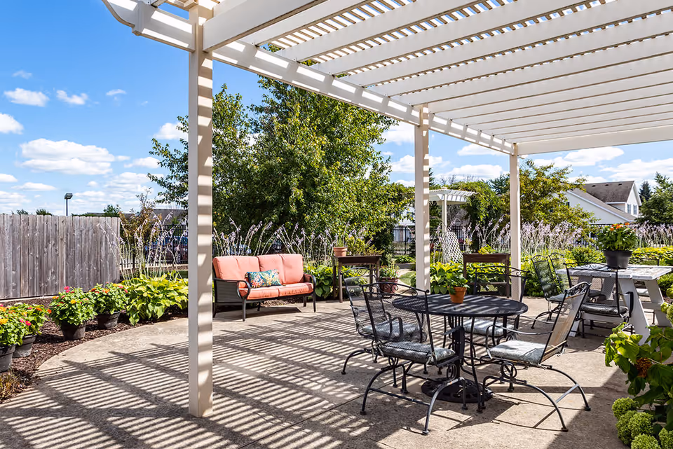 Outdoor patio area with a white pergola casting shadows on the concrete floor. The patio is furnished with black metal tables and chairs with cushions, a coral-colored cushioned bench, and several potted plants. Surrounding the patio are green bushes, flowering plants, and trees under a blue sky with scattered clouds.