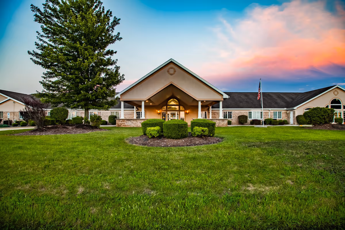 Front exterior view of Brookstone Estates of Mattoon South building with a large green lawn, trimmed bushes, a tall tree, and an American flagpole under a colorful sunset sky.
