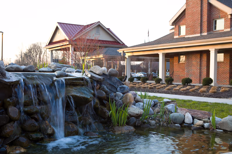 A small waterfall flowing over rocks into a pond with green plants around it, situated in front of a brick building with a covered porch and another building with a red roof in the background during daylight.