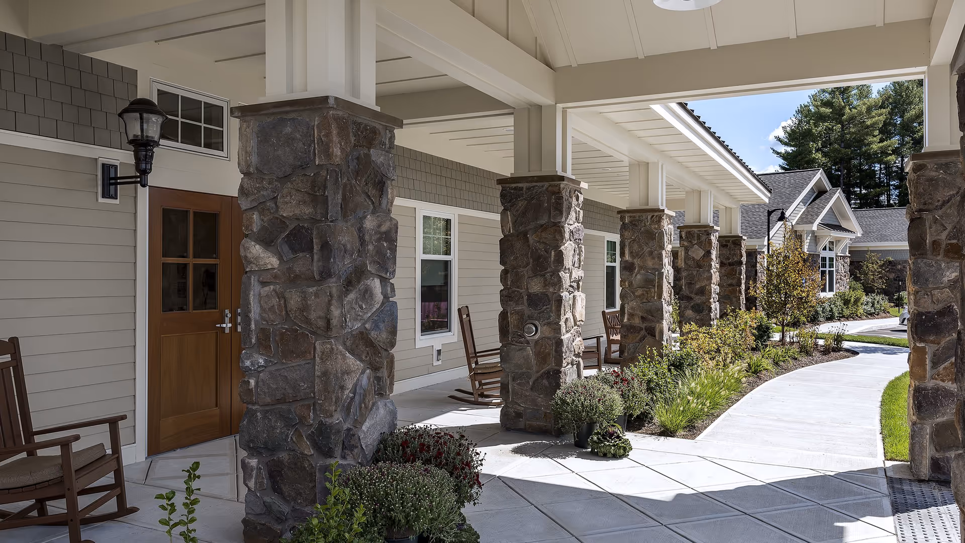 Covered outdoor walkway with stone pillars and wooden rocking chairs along the side of a building. There are potted plants and landscaping with bushes and small trees along a curved concrete path under a clear blue sky.