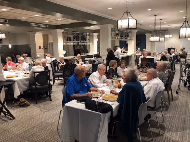 A dining room filled with elderly people seated at tables covered with white tablecloths, enjoying a meal together. The room has modern lighting fixtures, a carpeted floor, and a bar area in the background with glassware hanging above it.