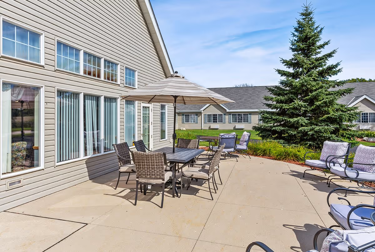 Outdoor patio area with a table and six chairs under a large umbrella, surrounded by additional cushioned chairs. The patio is adjacent to a beige building with multiple windows and a door. There is a large evergreen tree and well-maintained grass and landscaping in the background under a clear blue sky.