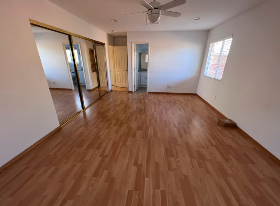 Empty bedroom with wood laminate floors, mirrored closet doors, a ceiling fan, and a window.