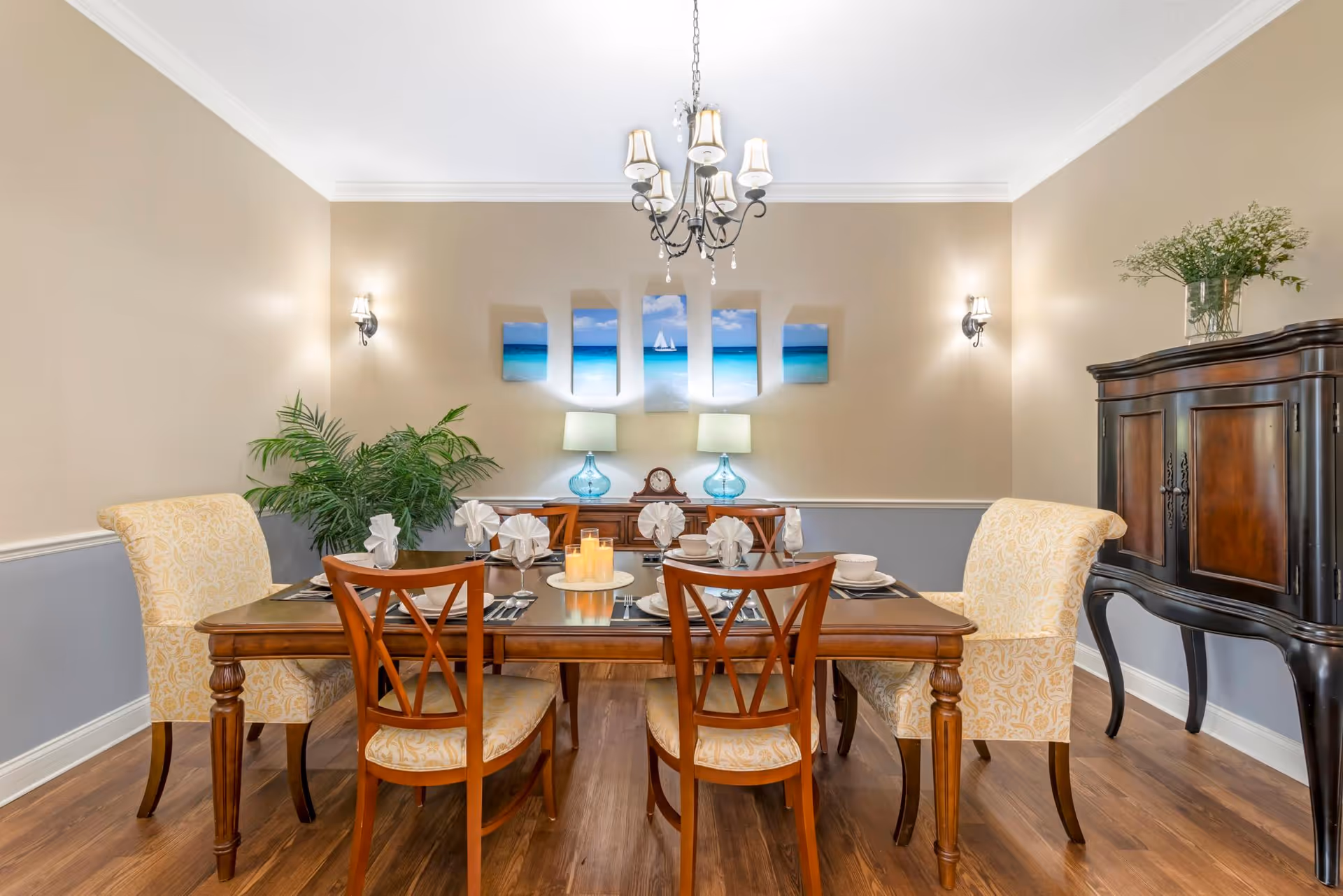 A well-lit dining room with a wooden dining table set for six, featuring two upholstered armchairs and four wooden chairs. The table is decorated with folded napkins, plates, and a centerpiece of candles. The room has beige and gray walls, a chandelier hanging from the ceiling, wall sconces, a large green plant in the corner, a wooden cabinet with a vase of flowers, and a five-panel artwork depicting a beach scene on the wall.