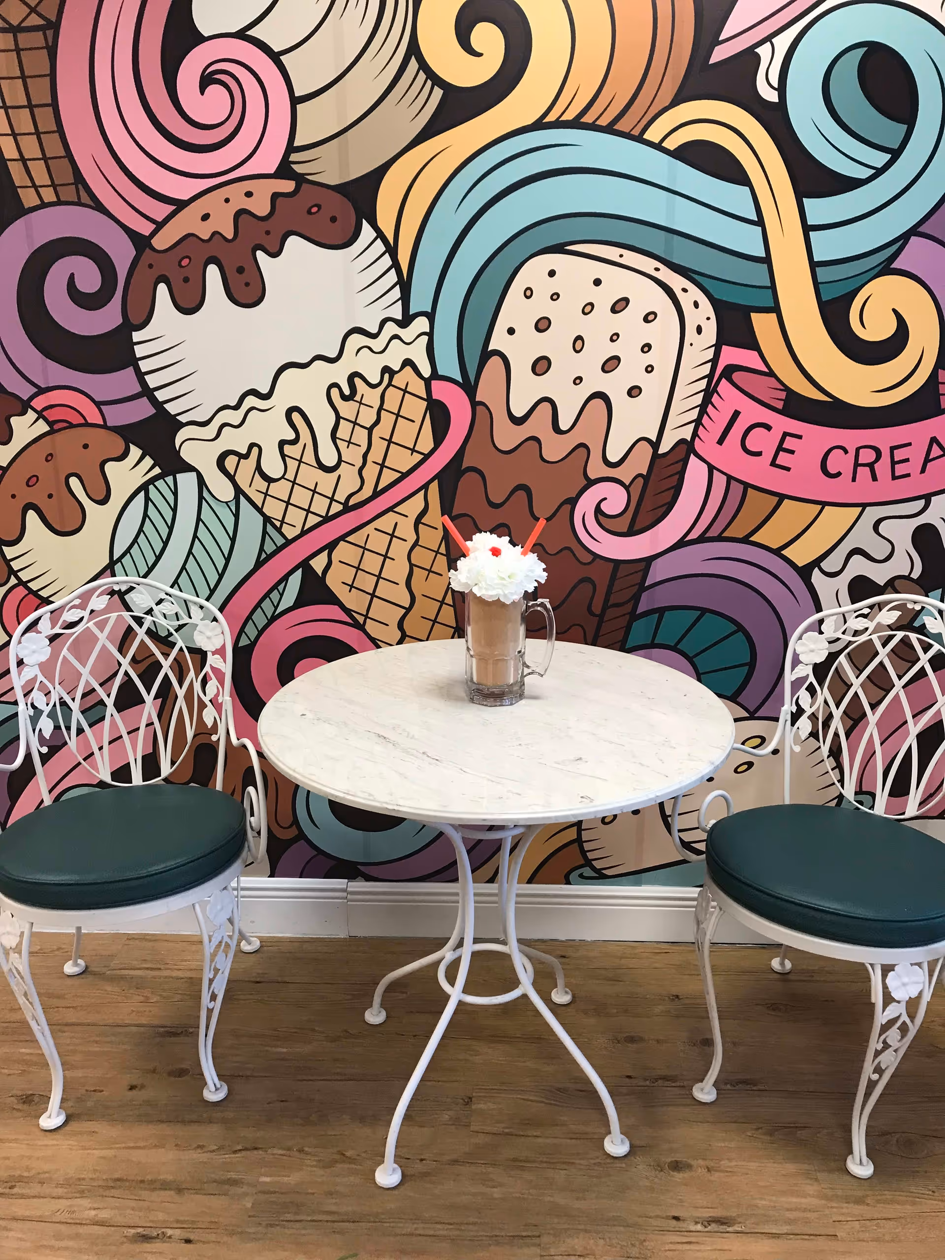 A small round white table with two white metal chairs with green cushions on a wooden floor. On the table is a glass mug filled with a chocolate milkshake topped with whipped cream and two red straws. The background features a colorful mural with large illustrations of ice cream cones, swirls, and the words 'ICE CREAM'.