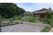 Paved courtyard with raised stone planters, wooden benches and a pergola beside a single-story building surrounded by greenery.