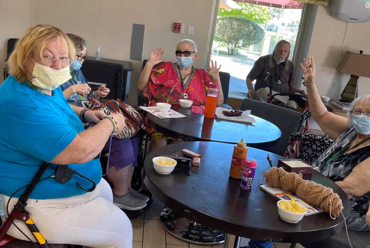 A group of elderly people sitting around two circular tables in a common area. Some are wearing face masks, and two individuals are raising their hands. On the tables are bowls of snacks, yarn, and other small items. A window in the background shows greenery outside.