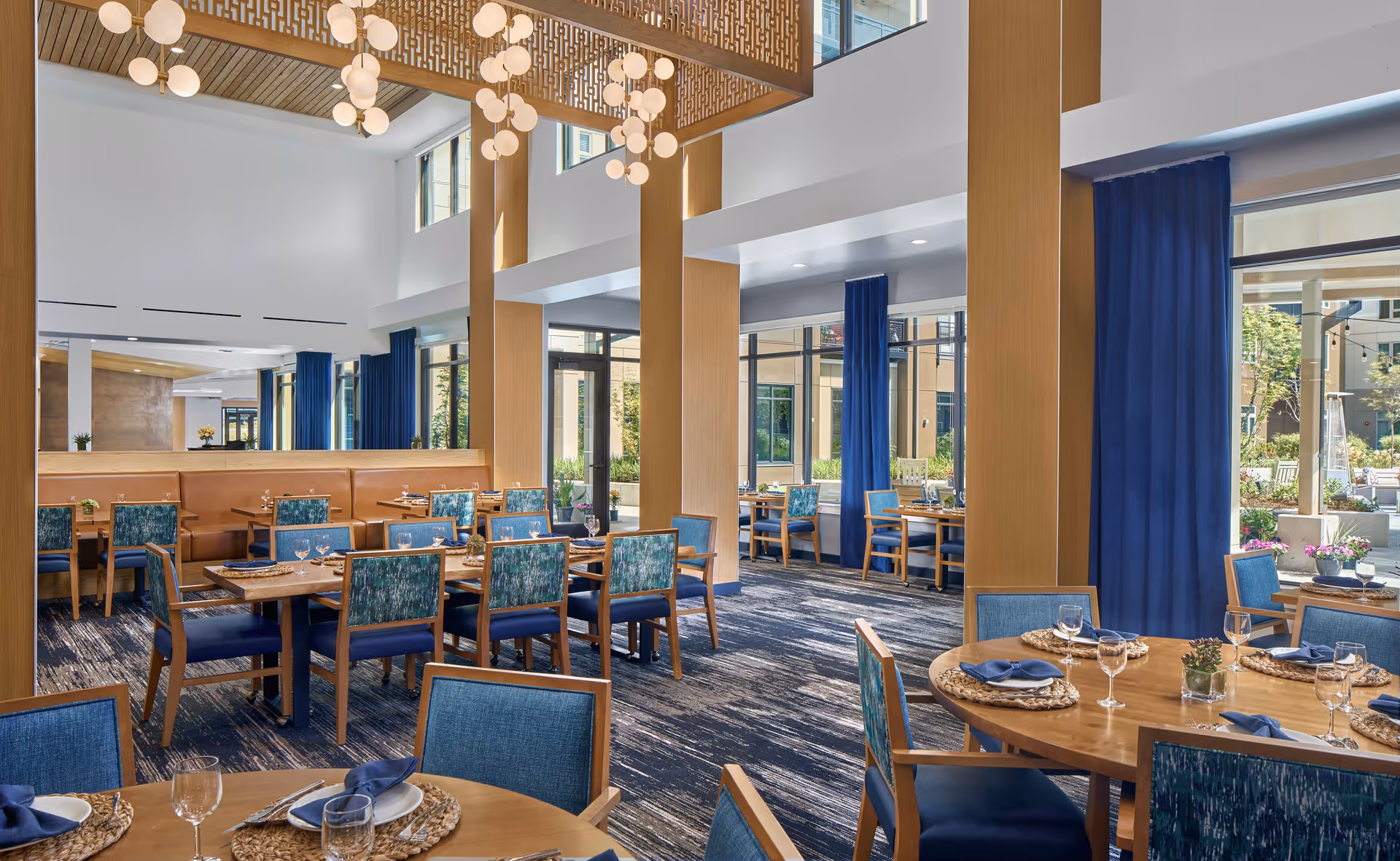 Bright and spacious dining room with wooden tables and blue cushioned chairs, set with plates, glasses, and napkins. Large windows with blue curtains allow natural light to fill the room, and modern light fixtures hang from the ceiling. The room features a mix of round and rectangular tables, with a view of an outdoor garden area.