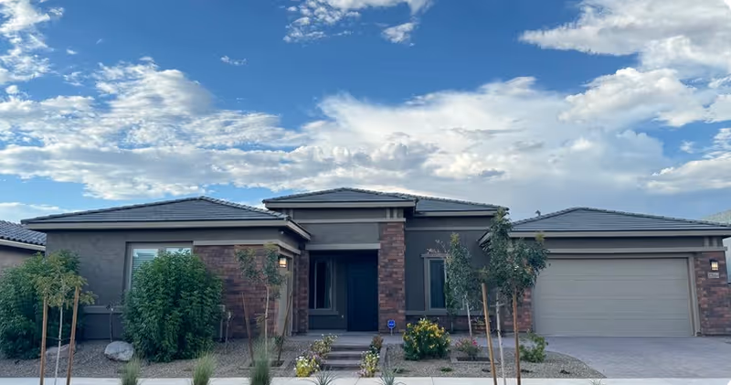 Single-story modern ranch-style building with a central entrance, attached garage, desert landscaping, and a blue sky with scattered clouds.