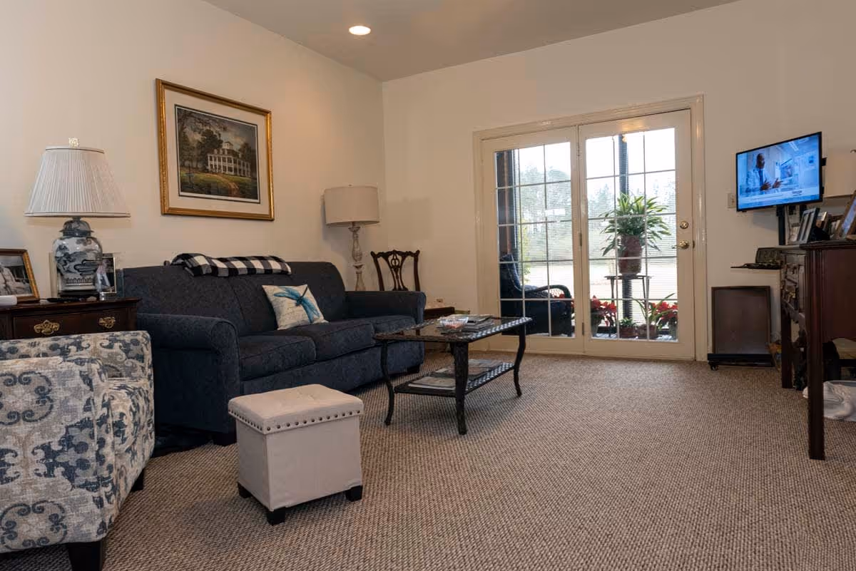 A cozy living room with a dark blue sofa, a patterned armchair, a small ottoman, and a glass coffee table. There are two lamps, a framed painting on the wall, and a TV mounted on the right wall. French doors lead to an outdoor area with plants visible through the glass.