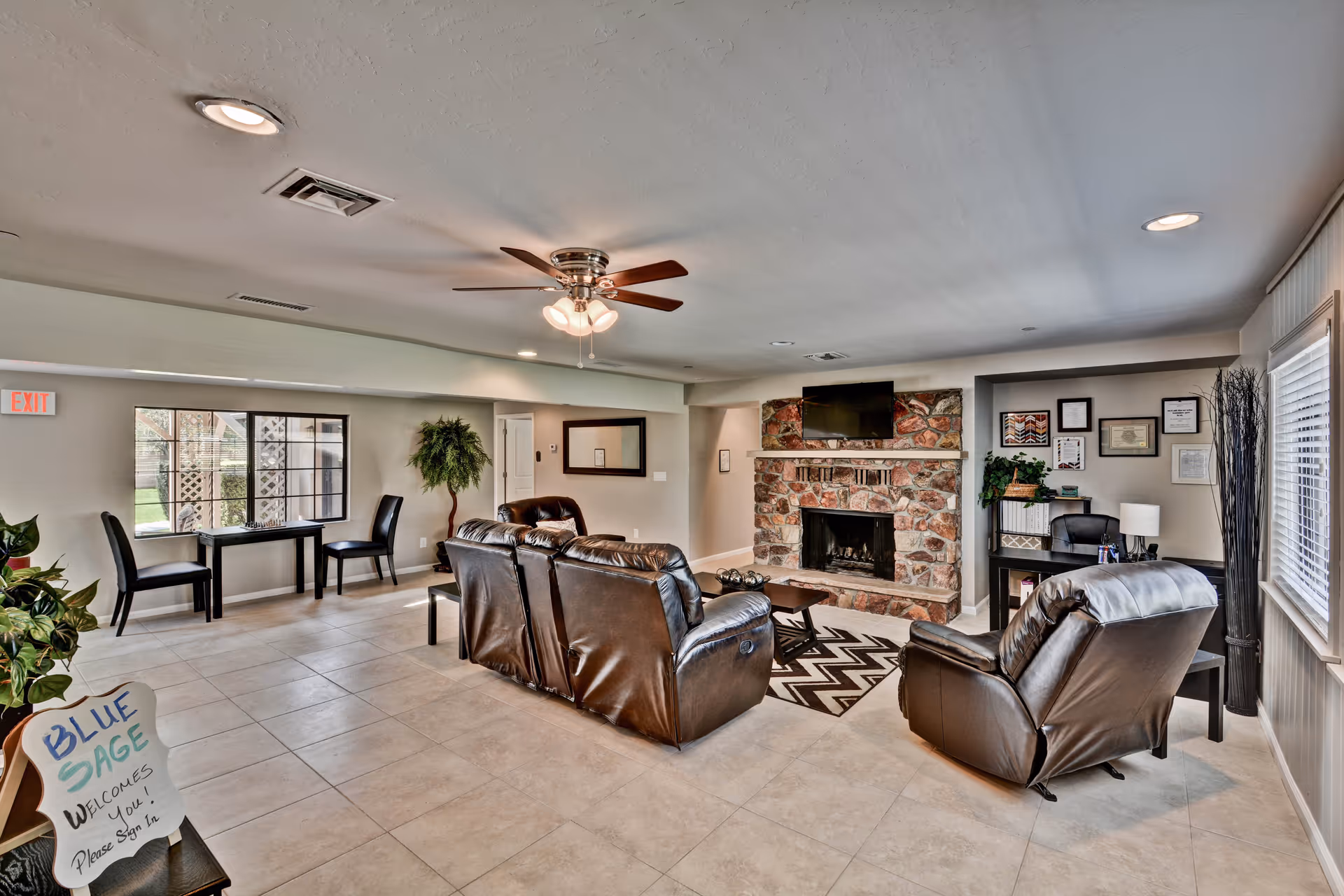 A spacious assisted living facility common area with beige tiled floors and light-colored walls. The room features a stone fireplace with a mounted TV above it, surrounded by a brown leather sofa set and a matching armchair on a patterned rug. There is a ceiling fan with lights in the center of the ceiling. Near the window, there is a small table with two chairs and a potted plant beside it. A sign on a small table near the entrance reads 'BLUE SAGE WELCOMES YOU! Please Sign In.' The room is well-lit with natural light coming through the windows and recessed ceiling lights.