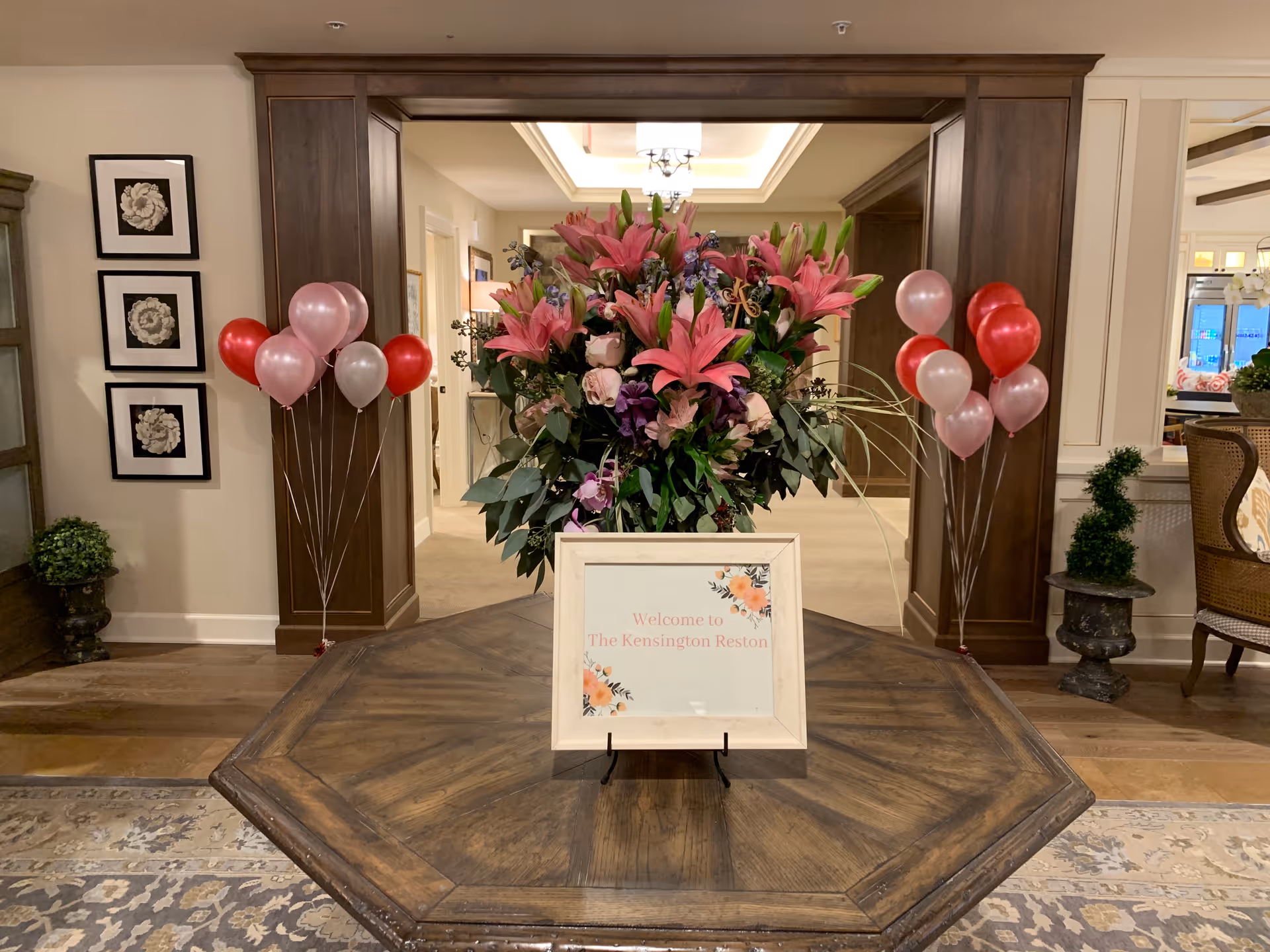 A wooden octagonal table with a large floral arrangement of pink lilies and other flowers in the center. In front of the flowers is a framed sign that reads 'Welcome to The Kensington Reston'. On either side of the table are clusters of pink, red, and silver balloons. The background shows a warmly lit interior with wood paneling, framed floral artwork on the wall, and decorative plants.