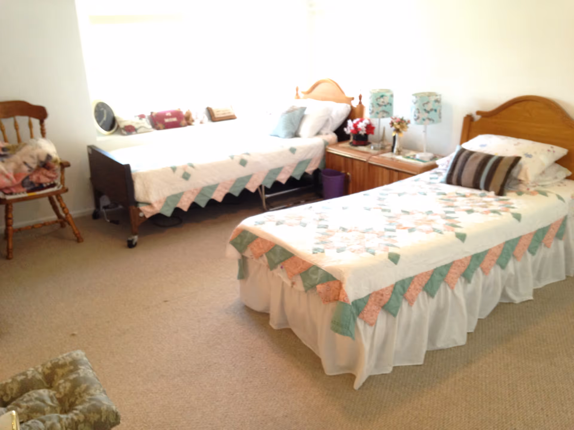 A bedroom with two single beds covered with patchwork quilts in pastel colors. Each bed has pillows and a wooden headboard. Between the beds is a wooden nightstand with two floral lamps and decorative items. There is a wooden chair with a folded blanket on it and a cushioned footstool on the carpeted floor.