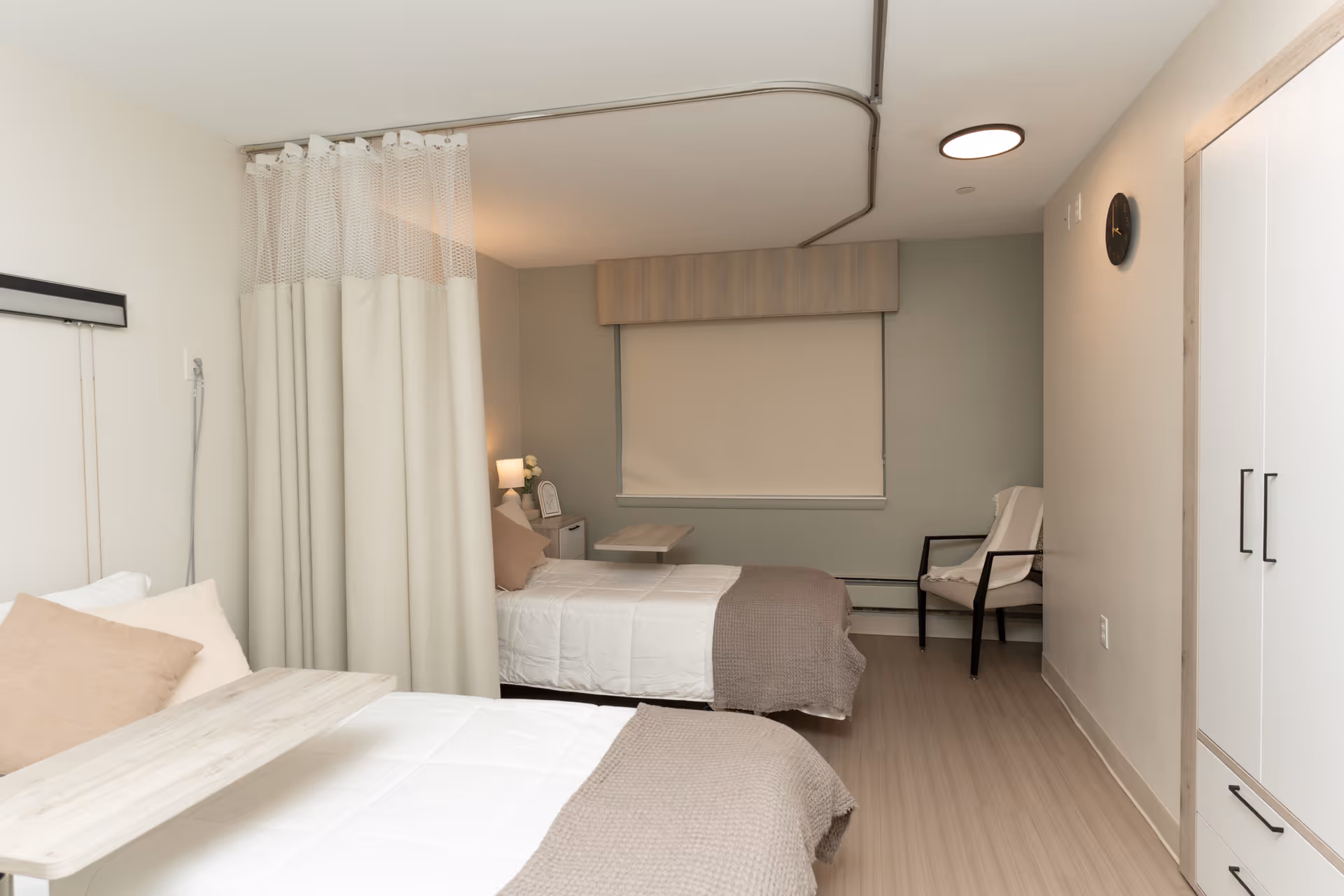A clean and simple bedroom with two single beds separated by a beige privacy curtain. Each bed has white bedding with beige throws and pillows. There is a small bedside table with a lamp and a clock between the beds. A white chair with black arms is placed near the window, which has a beige roller shade. A white wardrobe with black handles is on the right side of the room, and a round wall clock is mounted above it. The floor is light wood, and the walls are painted in neutral tones.
