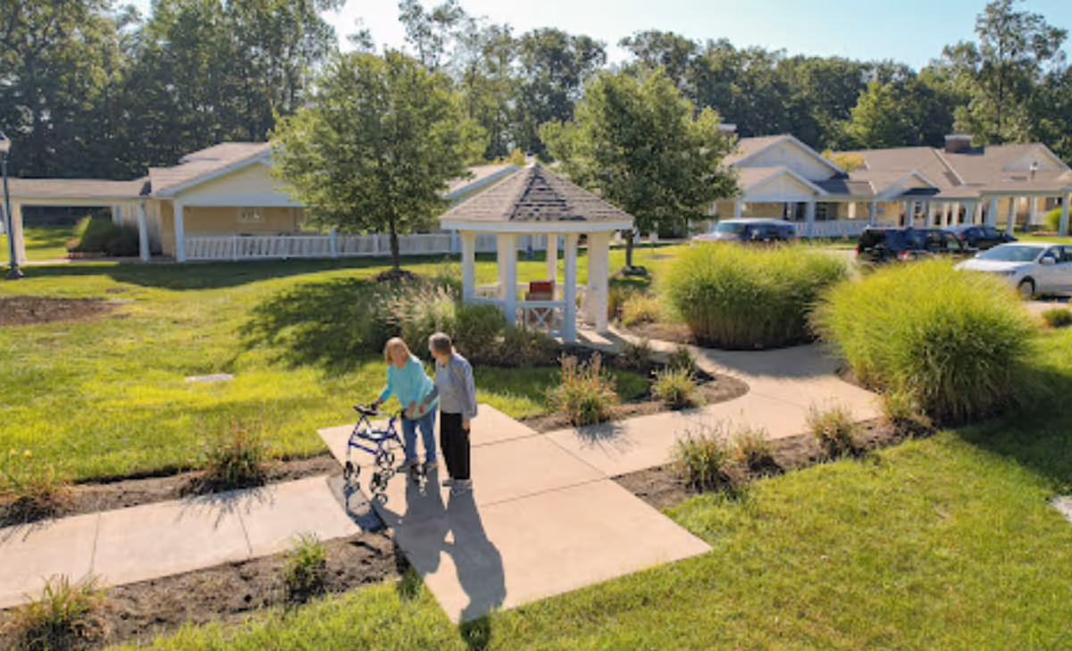 Two people using a walker on a paved path by a white gazebo and landscaped lawn in front of a single-story senior living building.