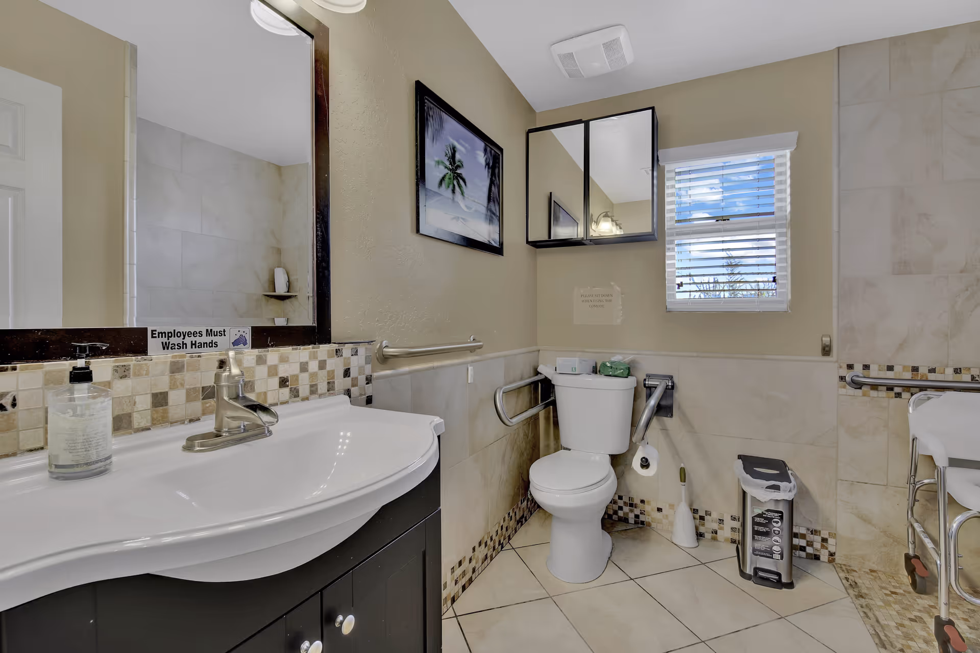 A clean and accessible bathroom with a white sink and silver faucet on a dark cabinet, a large mirror above the sink with a sign that reads 'Employees Must Wash Hands', a toilet with grab bars on both sides, a small window with blinds, a wall-mounted cabinet with mirrored doors, a trash can, and a shower area with a shower chair visible on the right side. The walls and floor are tiled in neutral tones, and there is a framed picture of palm trees on the wall.