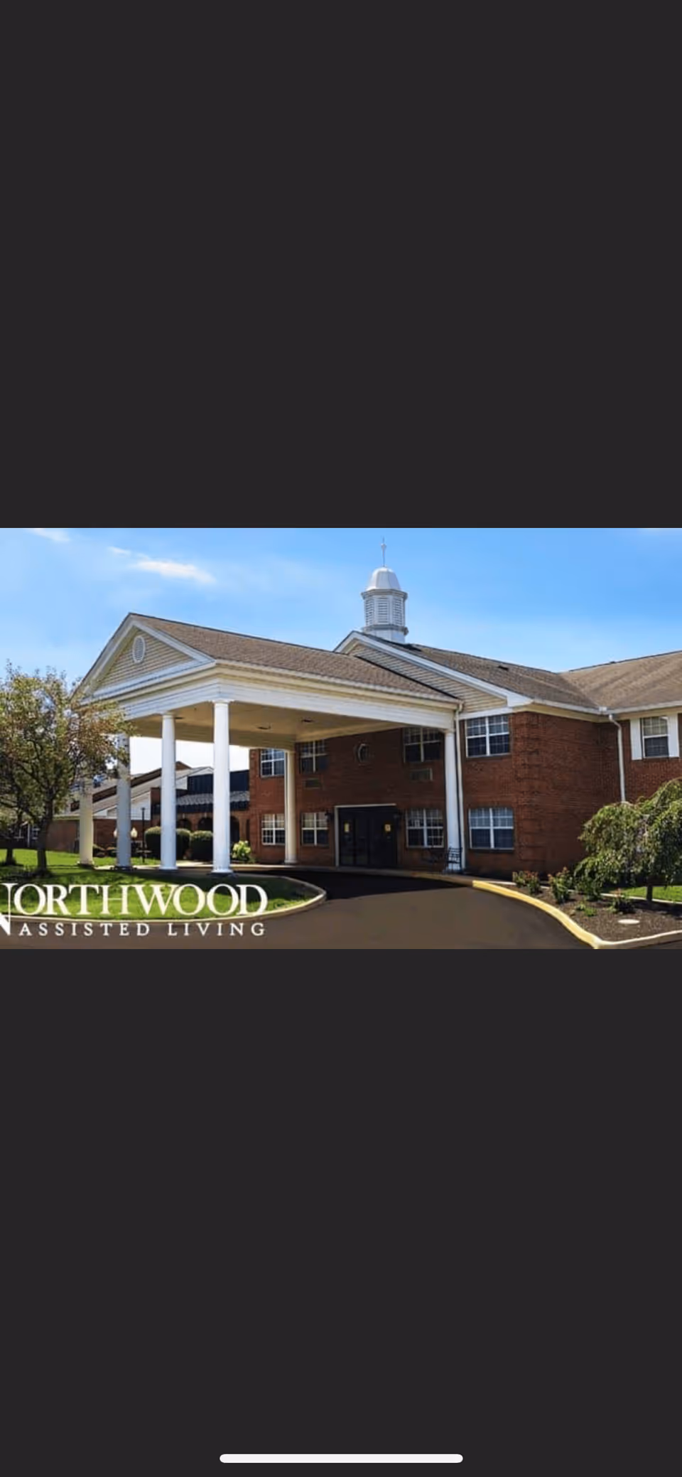 Exterior view of Northwood Assisted Living facility showing a brick building with white columns supporting a covered entrance, surrounded by green landscaping and a clear blue sky.