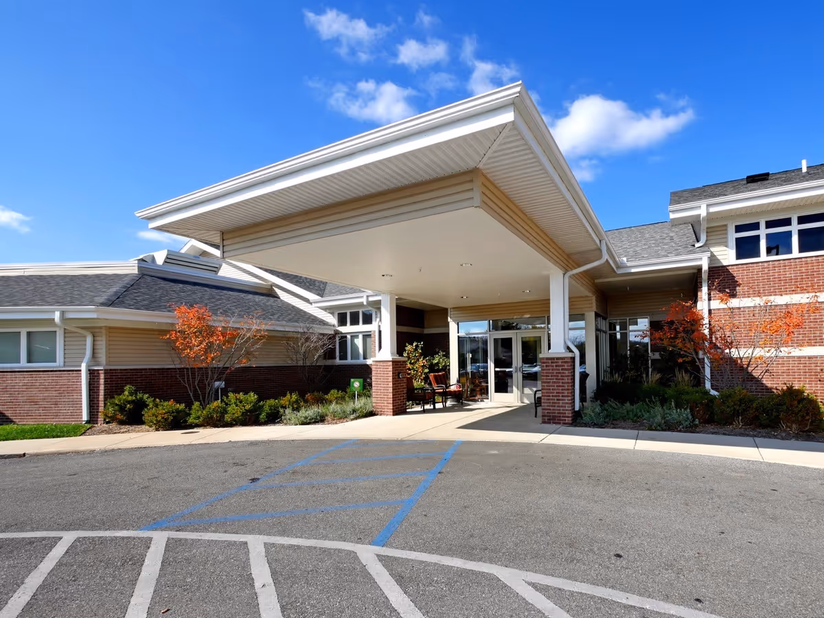 Entrance of a senior living facility with a covered drop-off area, brick and beige siding exterior, landscaped bushes and small trees, and a clear blue sky with a few clouds.