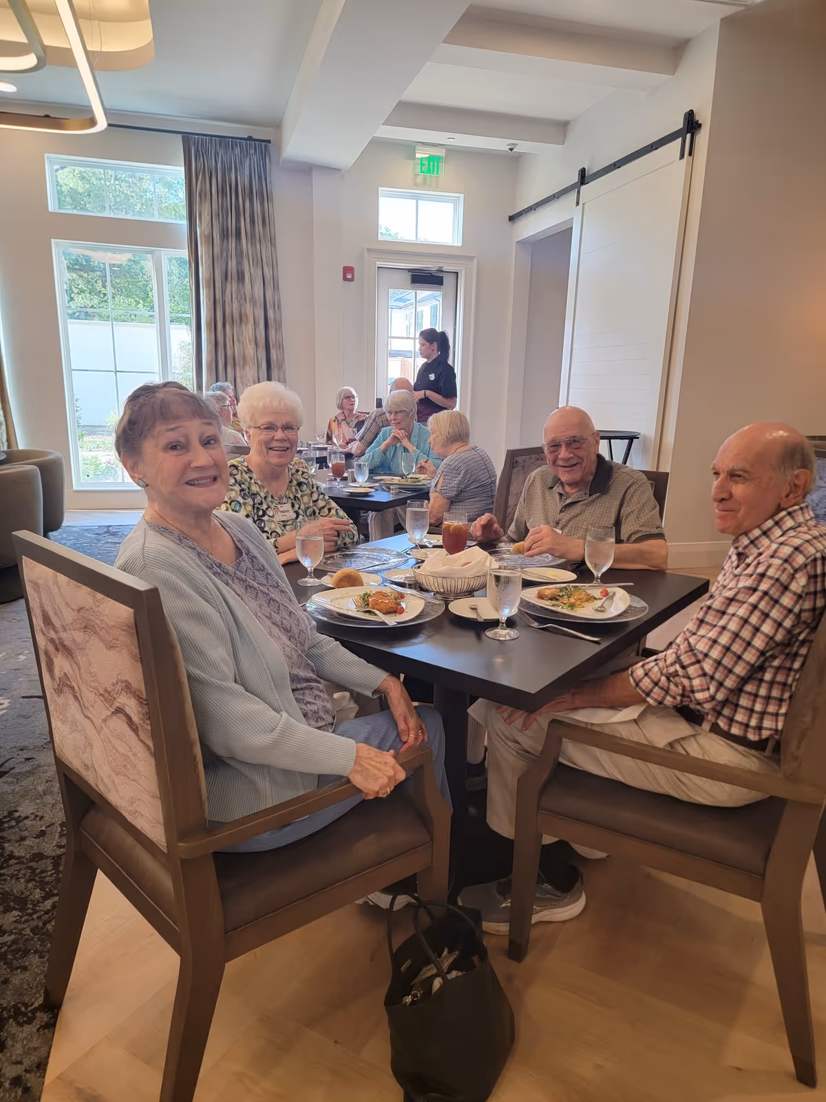A group of elderly people sitting around a dining table in a well-lit room, enjoying a meal together. There are plates with food, glasses of water, and a basket of bread on the table. In the background, more elderly individuals are seated at another table, and a staff member is standing near the door. The room has large windows with curtains and light-colored walls.