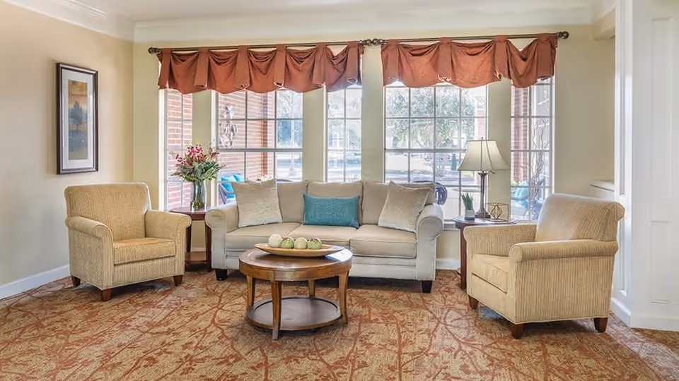 A cozy living room with a beige sofa adorned with three cushions, two beige armchairs, a round wooden coffee table with decorative items, a side table with a lamp and a small plant, and large windows with red valance curtains letting in natural light.