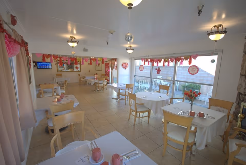 A bright dining room decorated with Valentine's Day themed decorations including red and pink garlands and heart-shaped ornaments hanging near the windows. The room has several tables covered with white tablecloths, each set with pink cups, saucers, and silverware. Light wooden chairs surround the tables, and large windows allow natural light to fill the space. The floor is tiled, and ceiling lights provide additional illumination.