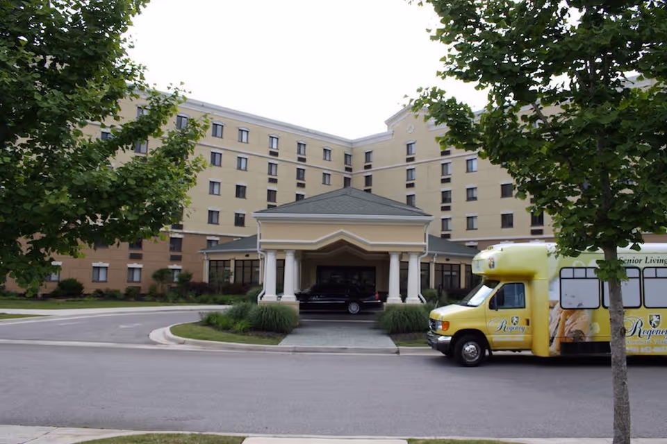Exterior view of a multi-story senior living facility with a covered entrance. A yellow shuttle bus with the Regency logo is parked on the right side, and a black car is under the entrance canopy. Trees and landscaping surround the driveway.