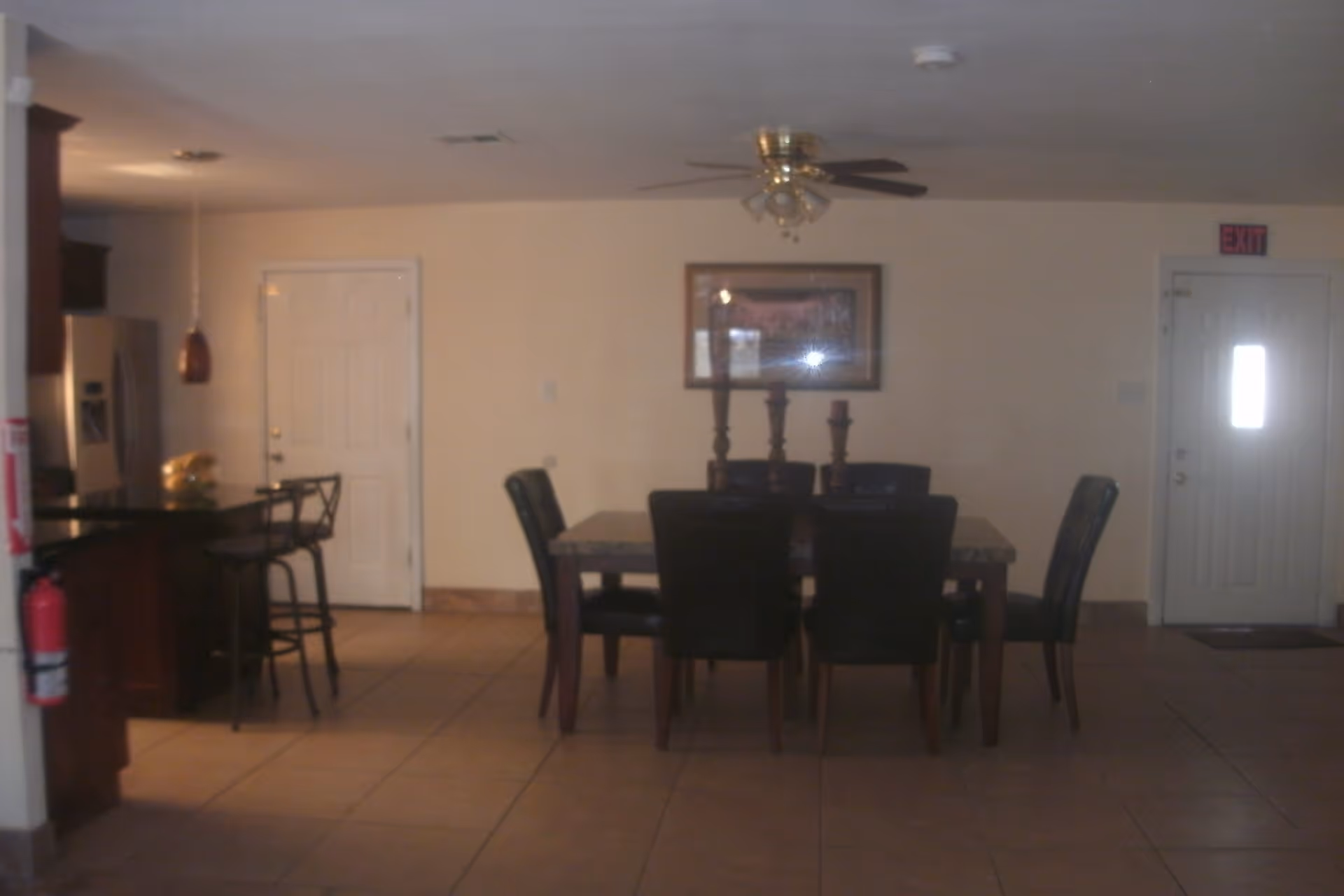 Interior view of a dining area with a rectangular dining table surrounded by six black chairs. On the table are three decorative candle holders. To the left, there is a kitchen counter with two bar stools and a hanging pendant light. A refrigerator is visible in the kitchen area. The floor is tiled, and the walls are light-colored. A ceiling fan with lights is mounted on the ceiling. There are two doors visible, one on the left side and one on the right side with an exit sign above it.