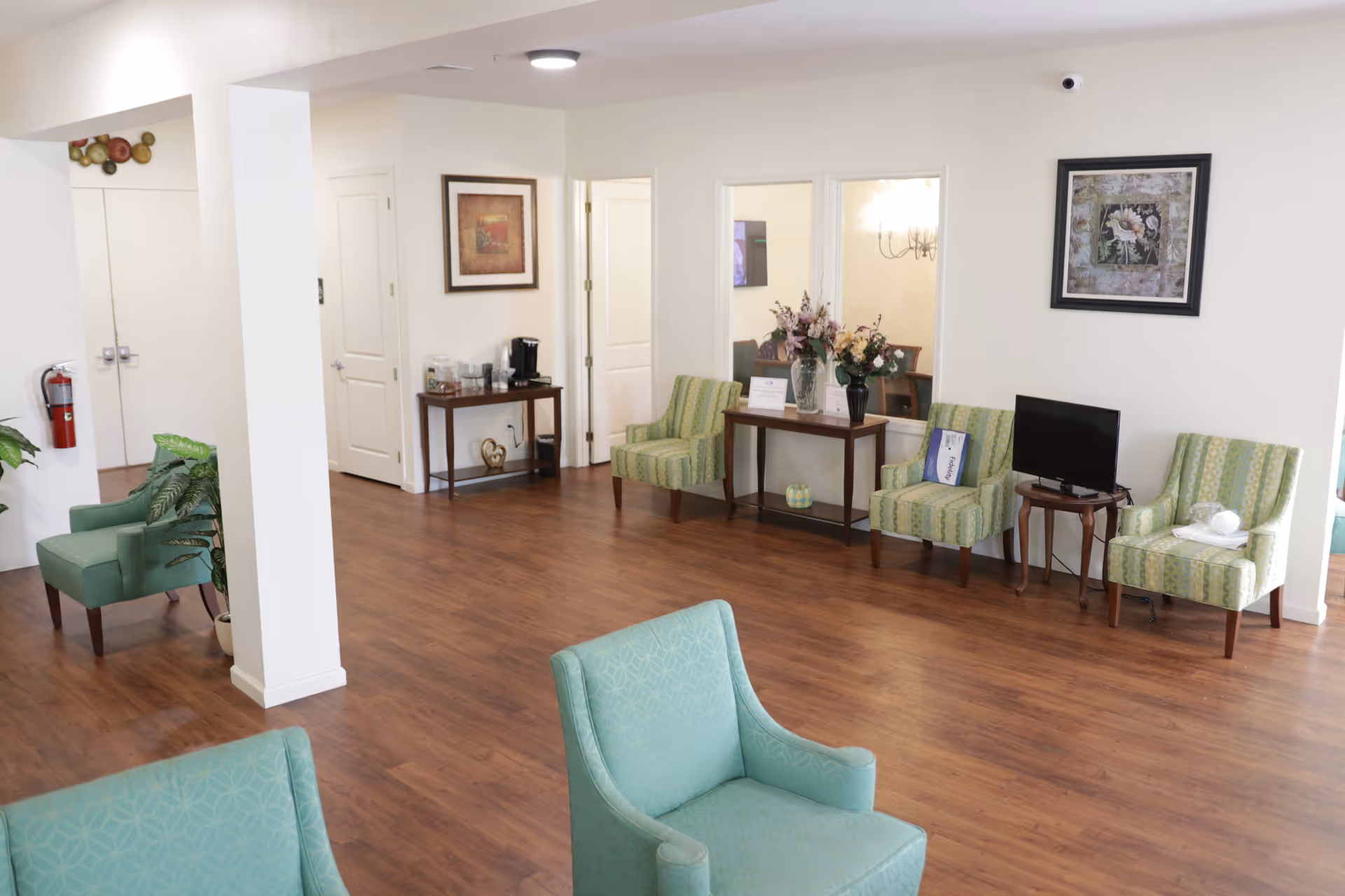 A spacious, well-lit common area with wooden flooring and light-colored walls. The room features several green upholstered chairs arranged along the walls and in the center. There are two small tables, one with a coffee maker and supplies, and the other with flower arrangements and a small TV. Wall art and a fire extinguisher are also visible.