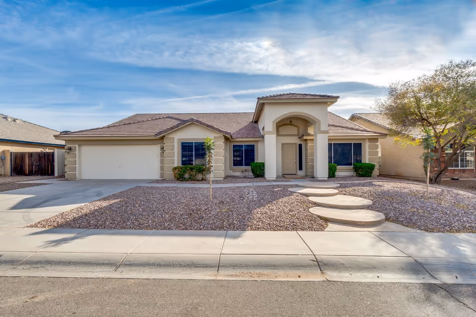 Front exterior view of a single-story house with a tiled roof, beige walls, a two-car garage, and a small landscaped yard with gravel and a few small trees under a partly cloudy sky.