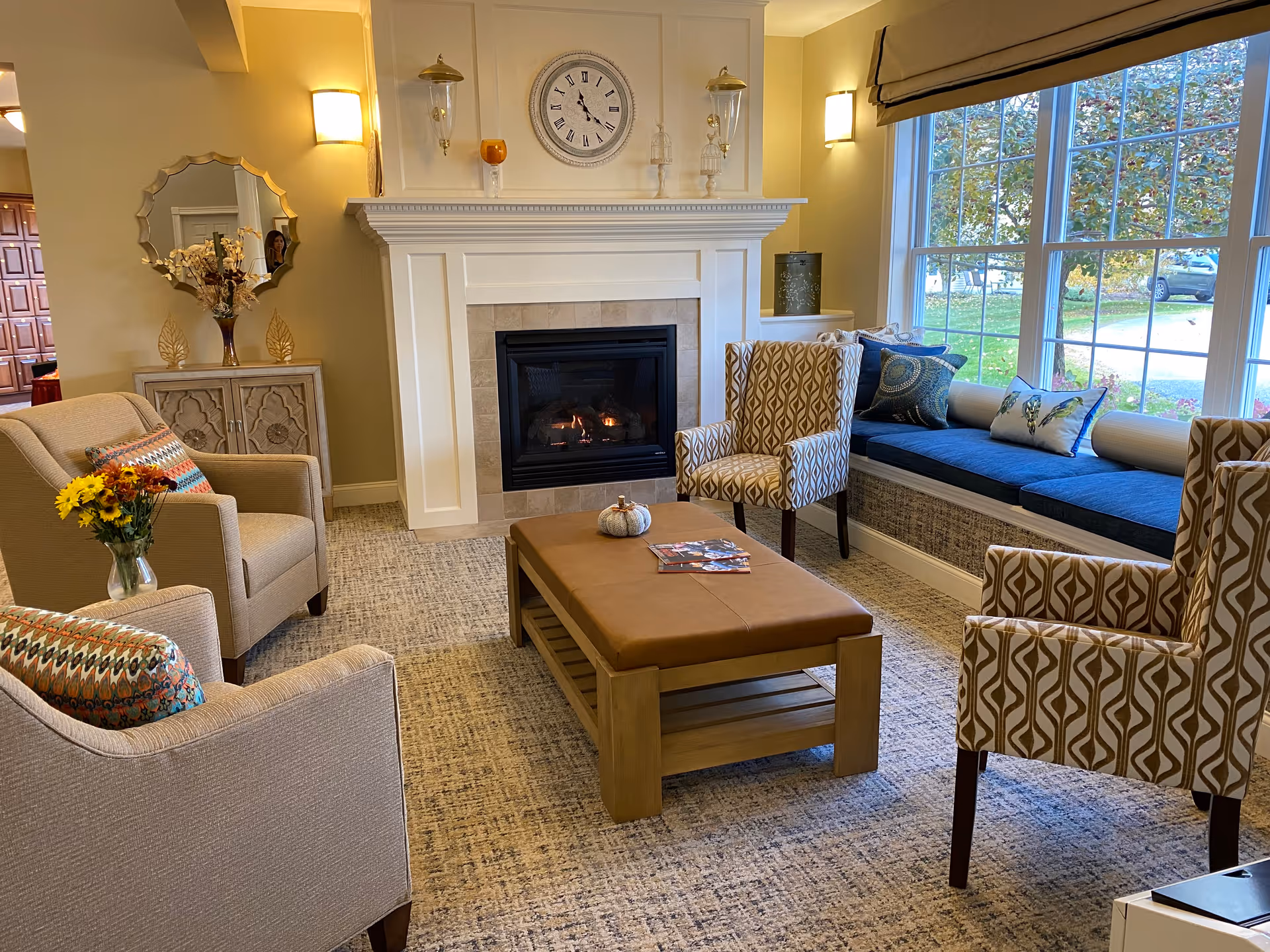 A cozy living room with a fireplace in the center, flanked by two wall sconces and decorative items on the mantle including a clock. The room features two beige armchairs with colorful cushions, two patterned armchairs, a wooden coffee table with a small pumpkin and magazines, and a window seat with blue cushions and pillows. Large windows show a view of green grass and trees outside.