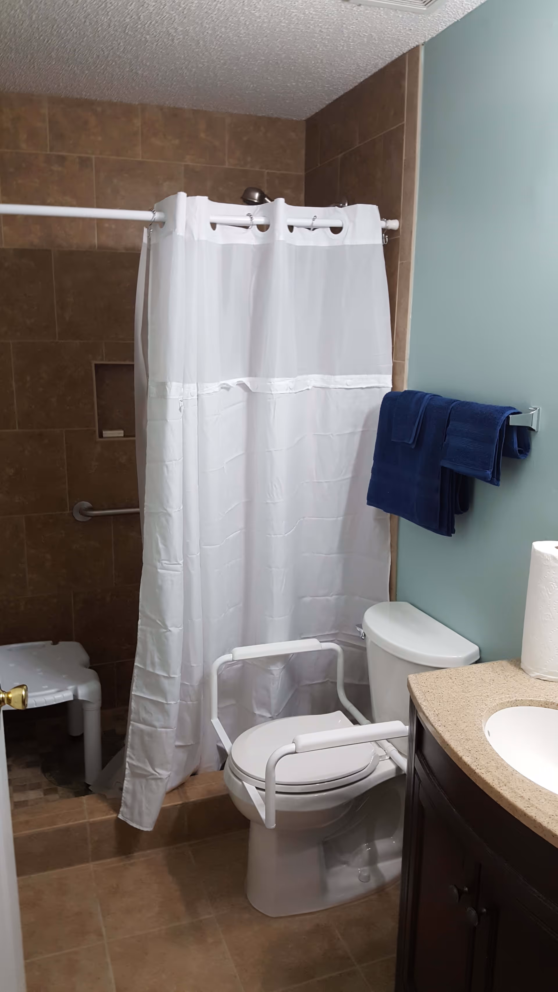 Accessible bathroom with a toilet fitted with support arms, a shower with a white curtain and bench, grab bars, towel on the wall, and a sink vanity.