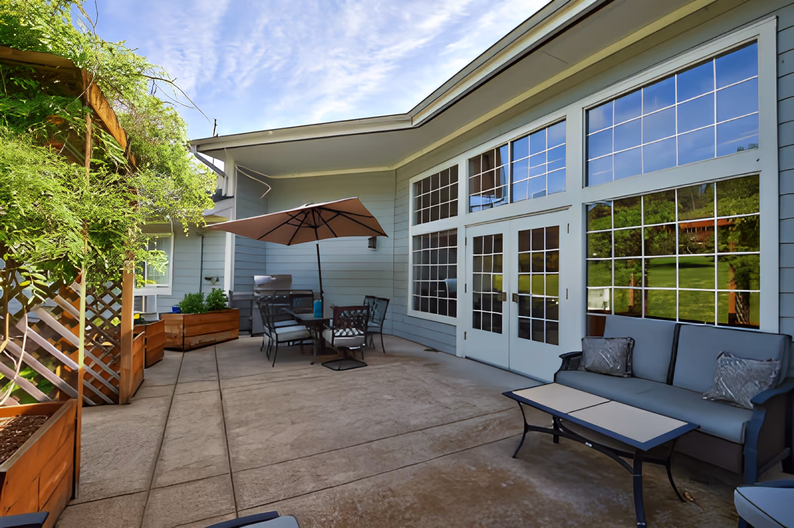 Outdoor patio area at Emerald Valley Assisted Living with a seating arrangement including a cushioned bench, chairs around a table with an umbrella, and large windows reflecting greenery and blue sky.