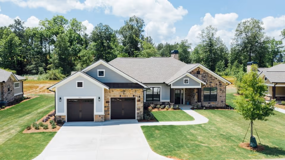 Front exterior view of a single-story house with a two-car garage, stone and siding facade, a concrete driveway, and a well-maintained lawn with a young tree. The house is surrounded by trees and under a partly cloudy sky.