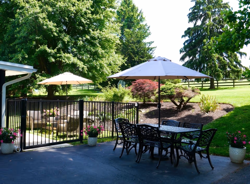 Outdoor patio area with a metal table and six chairs under a large beige umbrella. Behind a black metal fence, there is a seating area with wicker chairs and another umbrella. The patio is surrounded by green grass, trees, and landscaped bushes with flowers in pots near the fence.