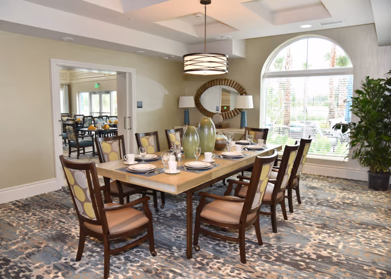 A well-lit dining room with a rectangular wooden table set for eight people. The table has plates, cups, glasses, and decorative vases. The room features a large arched window with blinds, a round mirror on the wall, two table lamps, and a patterned carpet. Through an open doorway, another dining area with tables and chairs is visible.