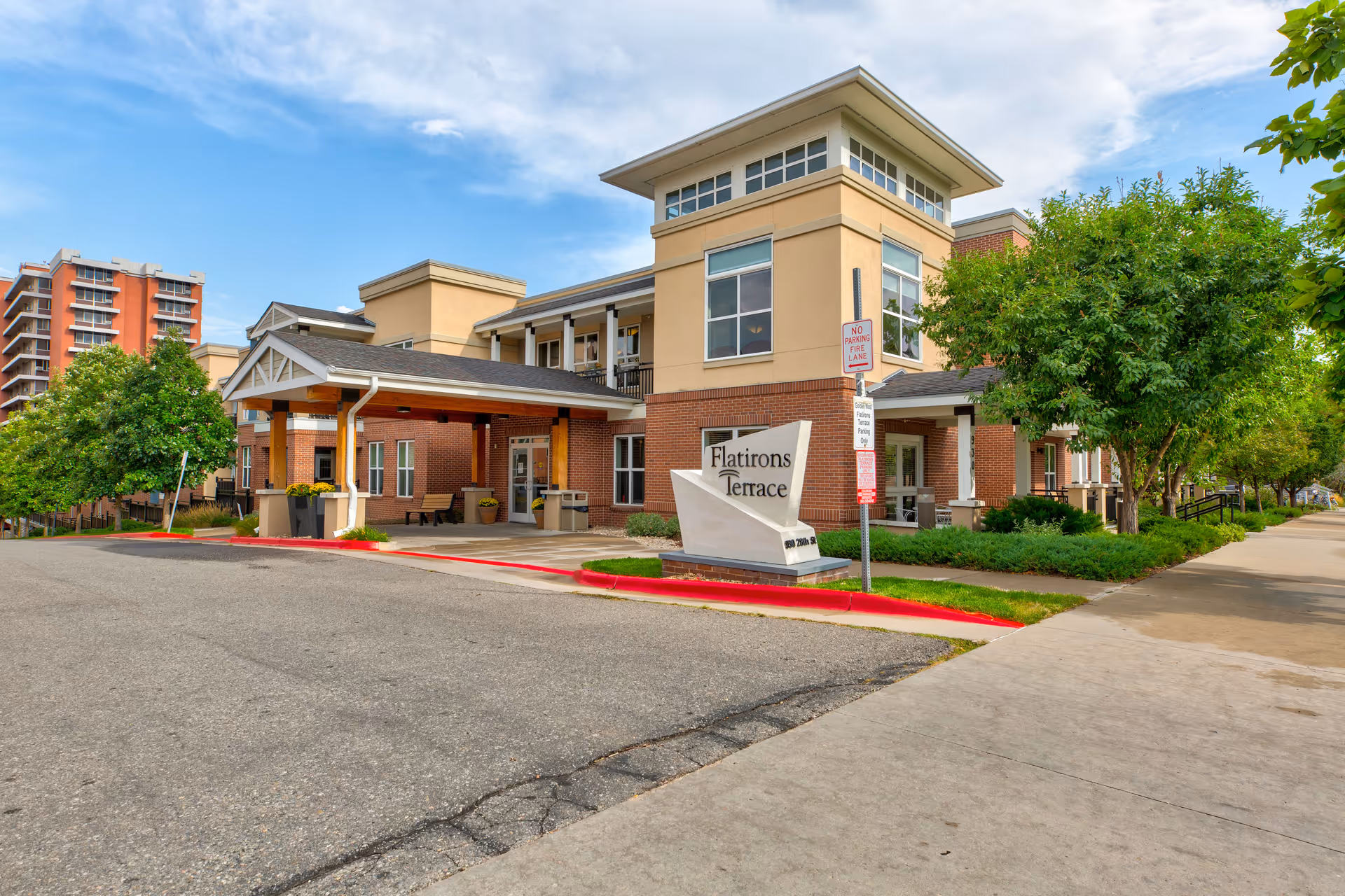 Exterior view of Flatirons Terrace Senior Living facility showing a modern two-story building with a covered entrance, surrounded by trees and landscaping under a partly cloudy sky.