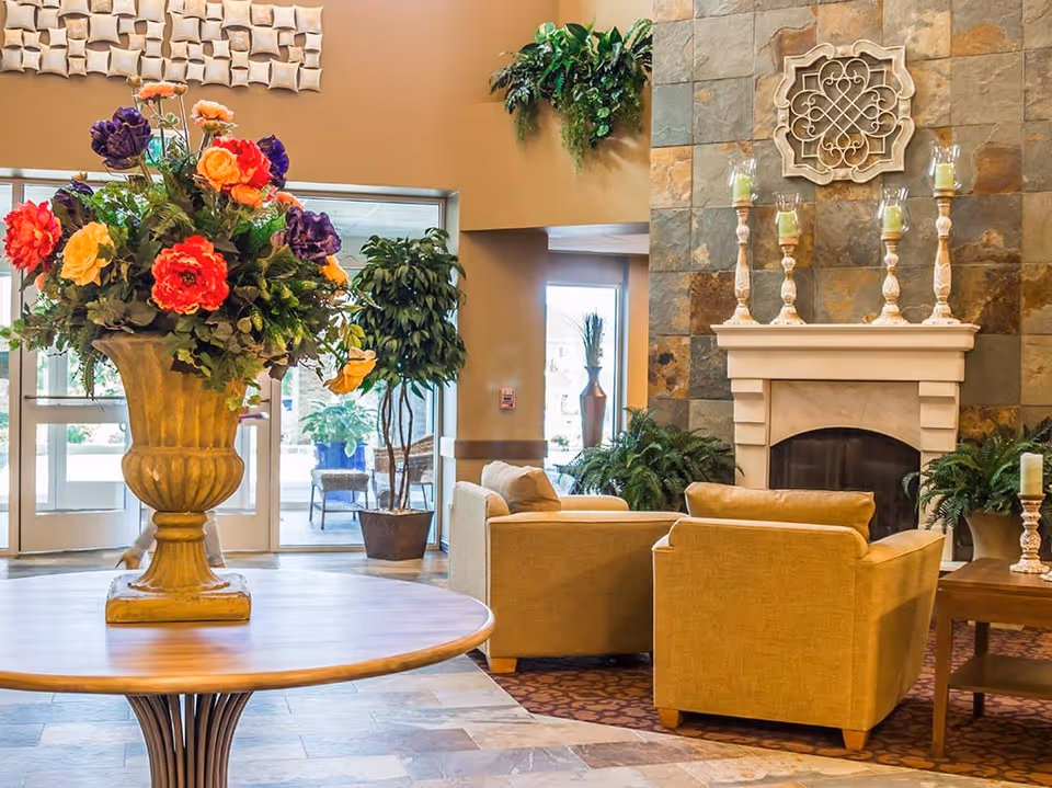 A cozy living room area with two beige armchairs facing a stone fireplace adorned with five candle holders. A large vase with colorful flowers sits on a round wooden table in the foreground. There are green plants around the room and large windows letting in natural light.