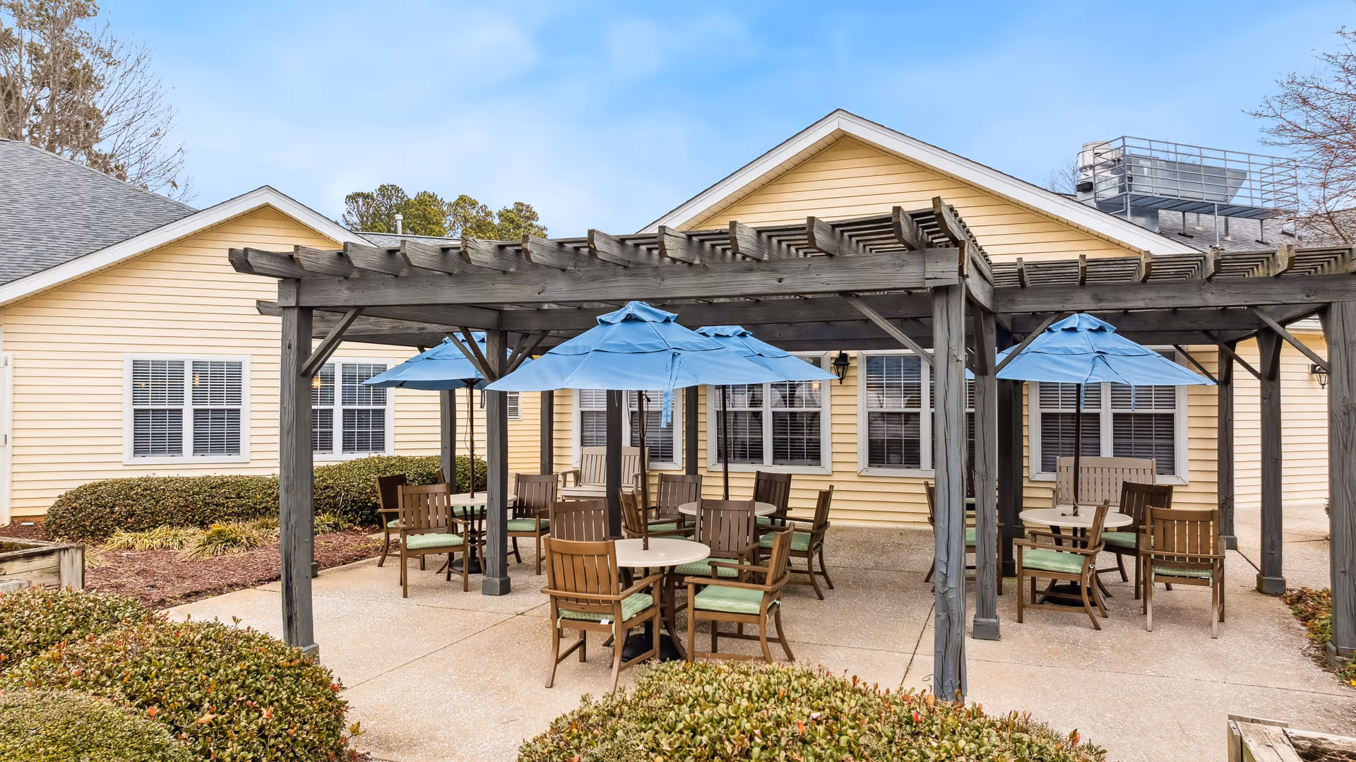 Outdoor patio area with several round tables and wooden chairs with green cushions under blue umbrellas, shaded by a wooden pergola. The patio is adjacent to a yellow building with white-trimmed windows and surrounded by neatly trimmed bushes.