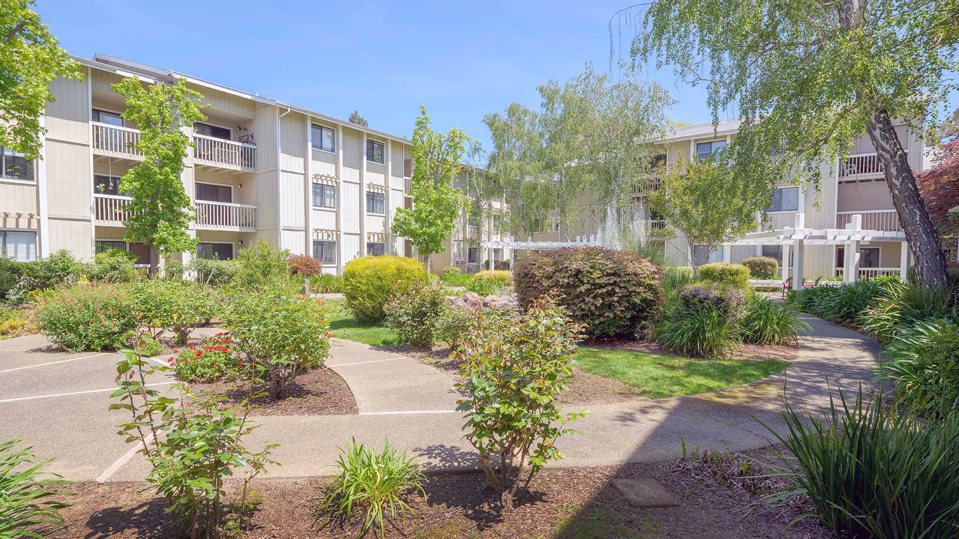 Outdoor courtyard area of a senior living facility with paved walkways, green bushes, trees, and a three-story building in the background under a clear blue sky.
