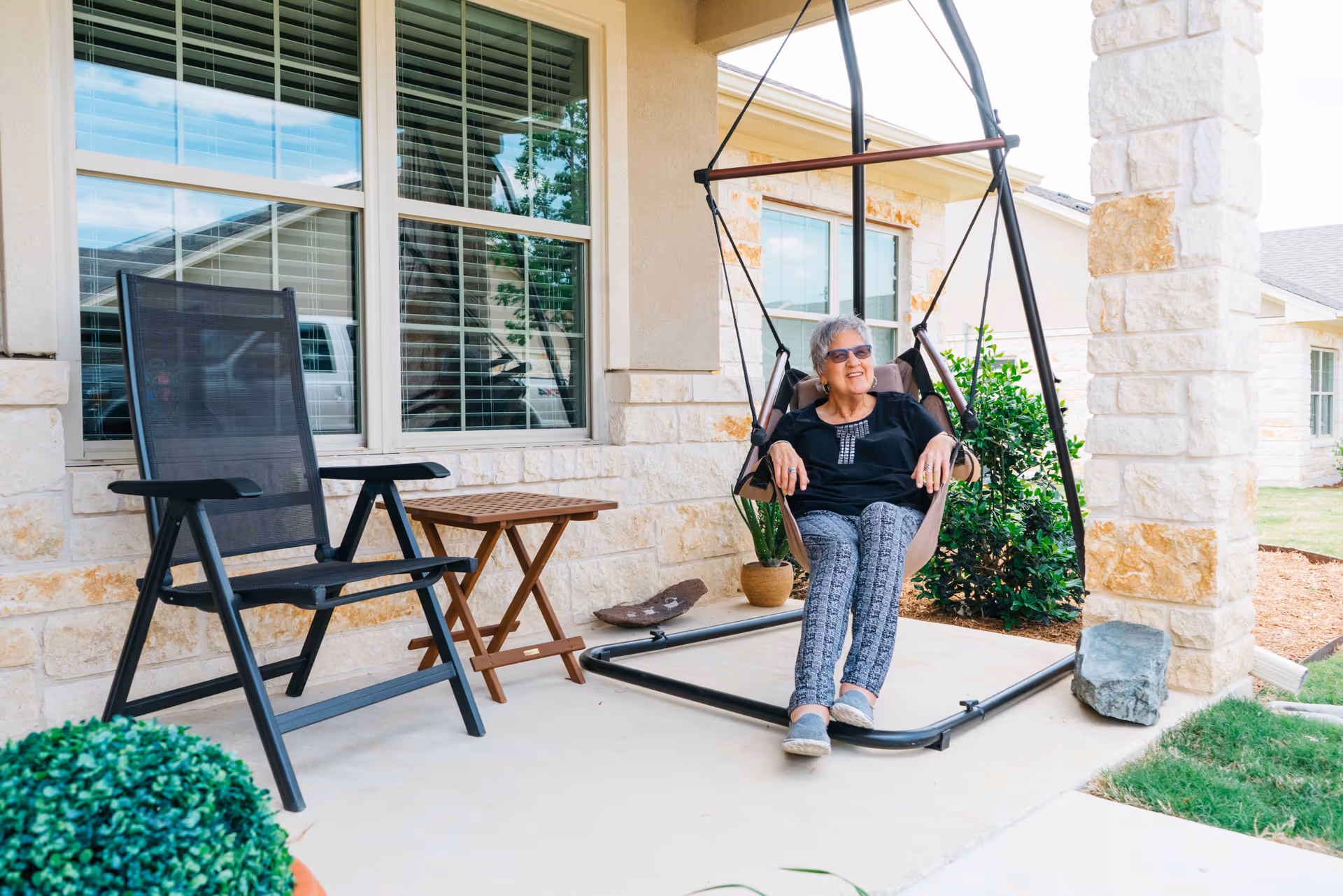 An elderly woman with short gray hair and sunglasses is sitting and relaxing on a hanging chair on a covered porch. Next to her is a black mesh chair and a small wooden table. The porch is part of a stone and stucco building with large windows and some greenery around.