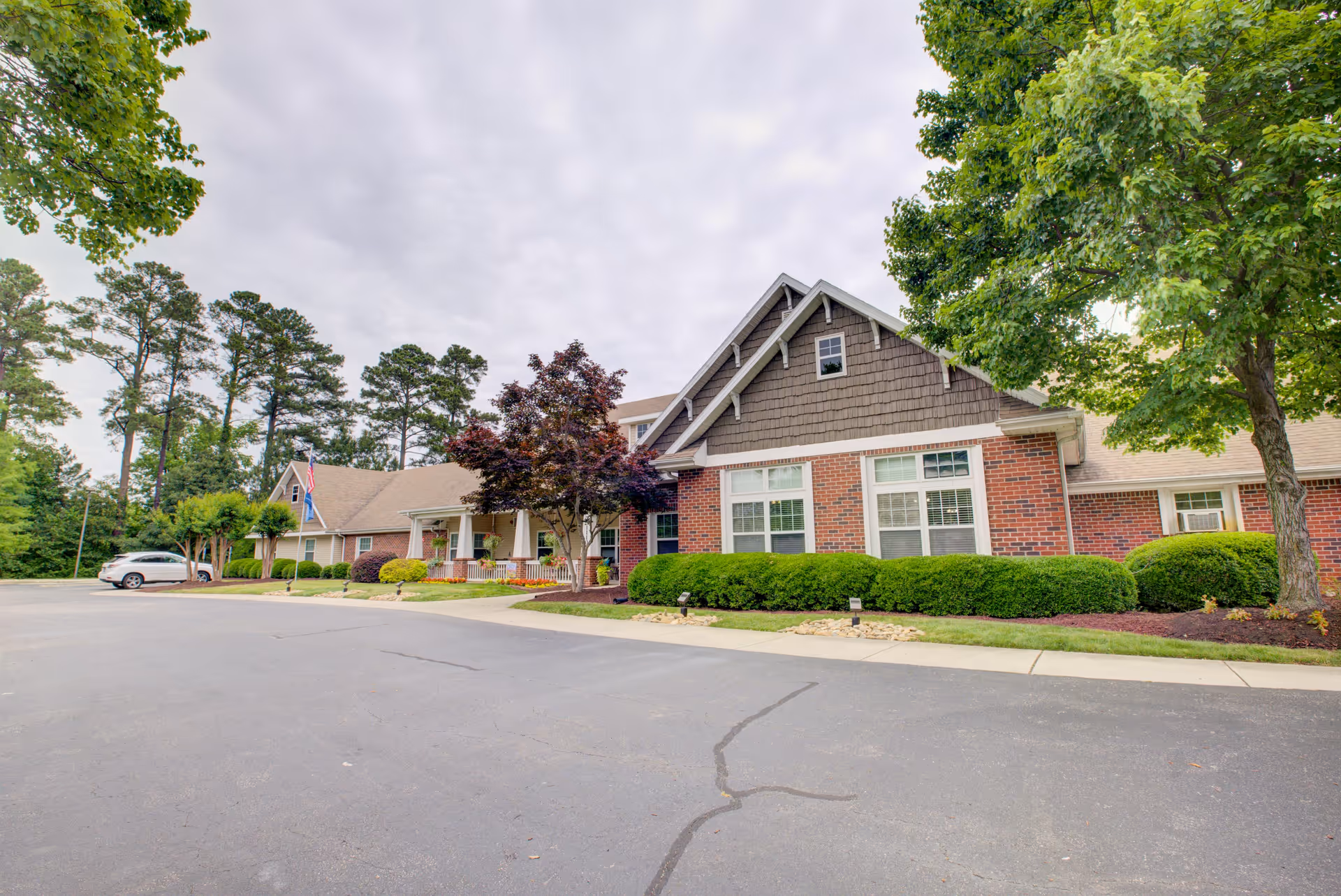 Exterior view of Falls River Village Assisted Living facility showing a single-story brick building with a gabled roof, surrounded by well-maintained landscaping including bushes and trees, with a paved driveway in front and a cloudy sky overhead.
