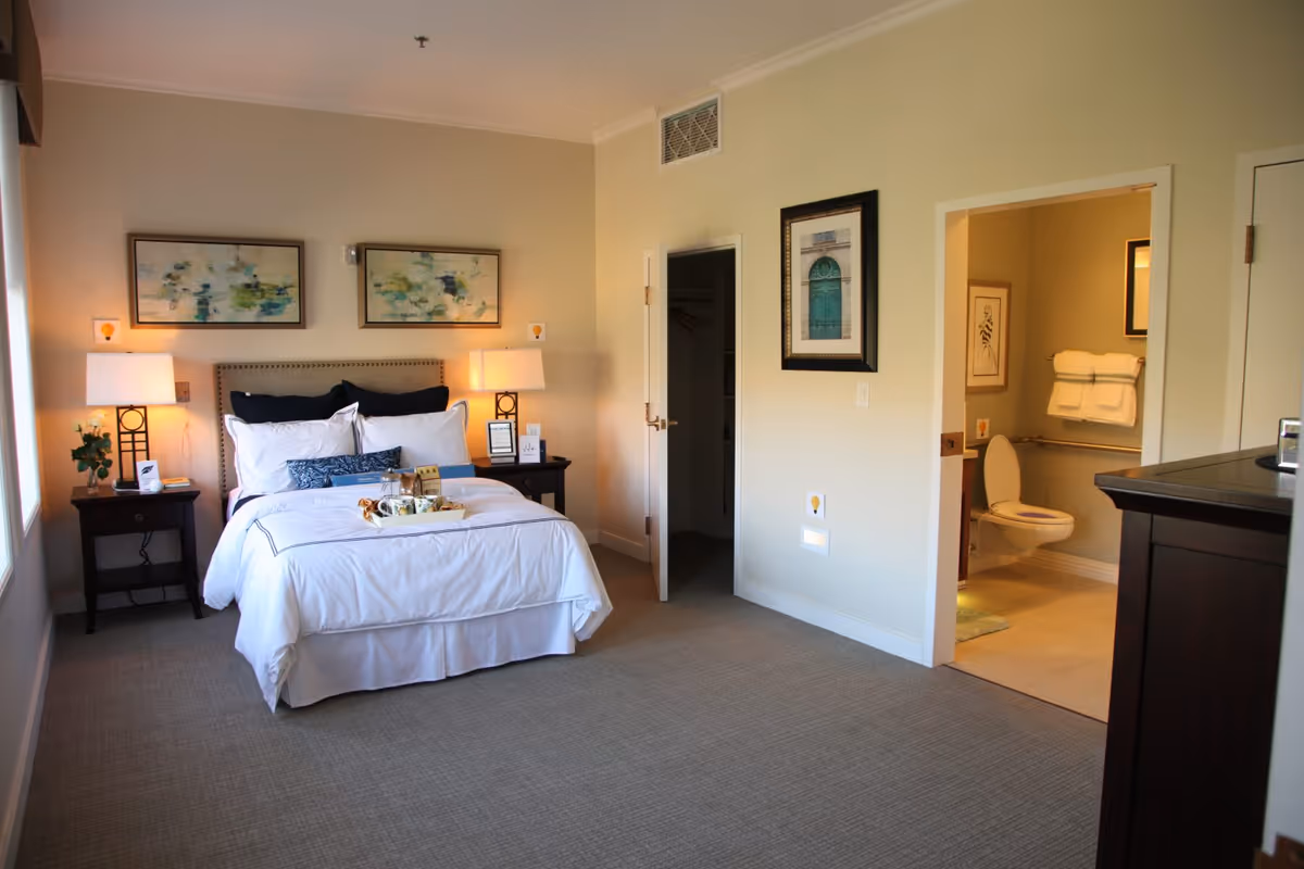 A neatly made bed with white linens and decorative pillows in a bedroom. Two bedside tables with lamps flank the bed. Two abstract paintings hang above the bed. To the right, an open door reveals a bathroom with a toilet, towels, and framed artwork. Another door leads to a closet. The room has beige walls and carpeted floor.